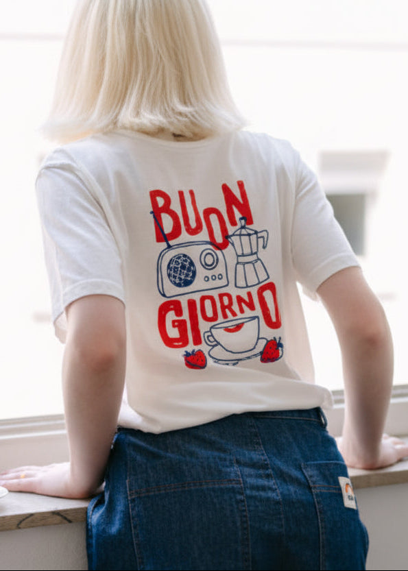 Person wearing a t-shirt with 'Buon Giorno' and coffee-themed design, standing by a window.